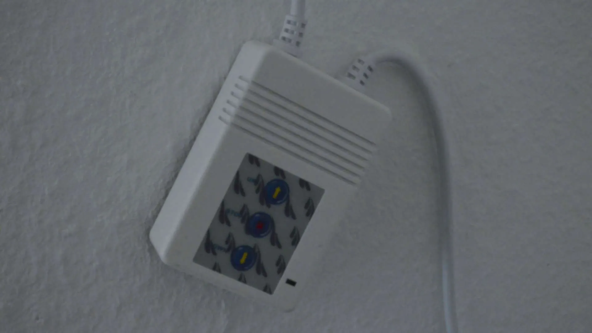 The original projector screen controller mounted on a white textured wall: a white plastic box with three round blue push buttons arranged vertically, each marked with a yellow indicator, connected by white cables.