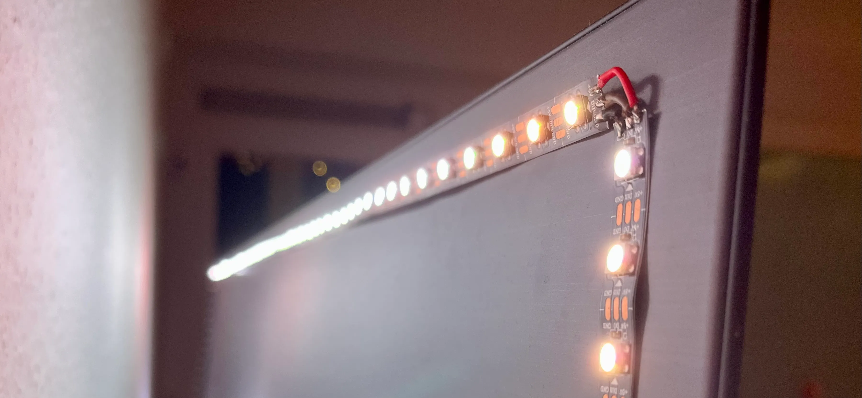 Close-up side view of an addressable LED strip adhered along the top and right edges of the back of a TV panel, with individual LEDs lit warm white and a red power wire connected at the top-right corner