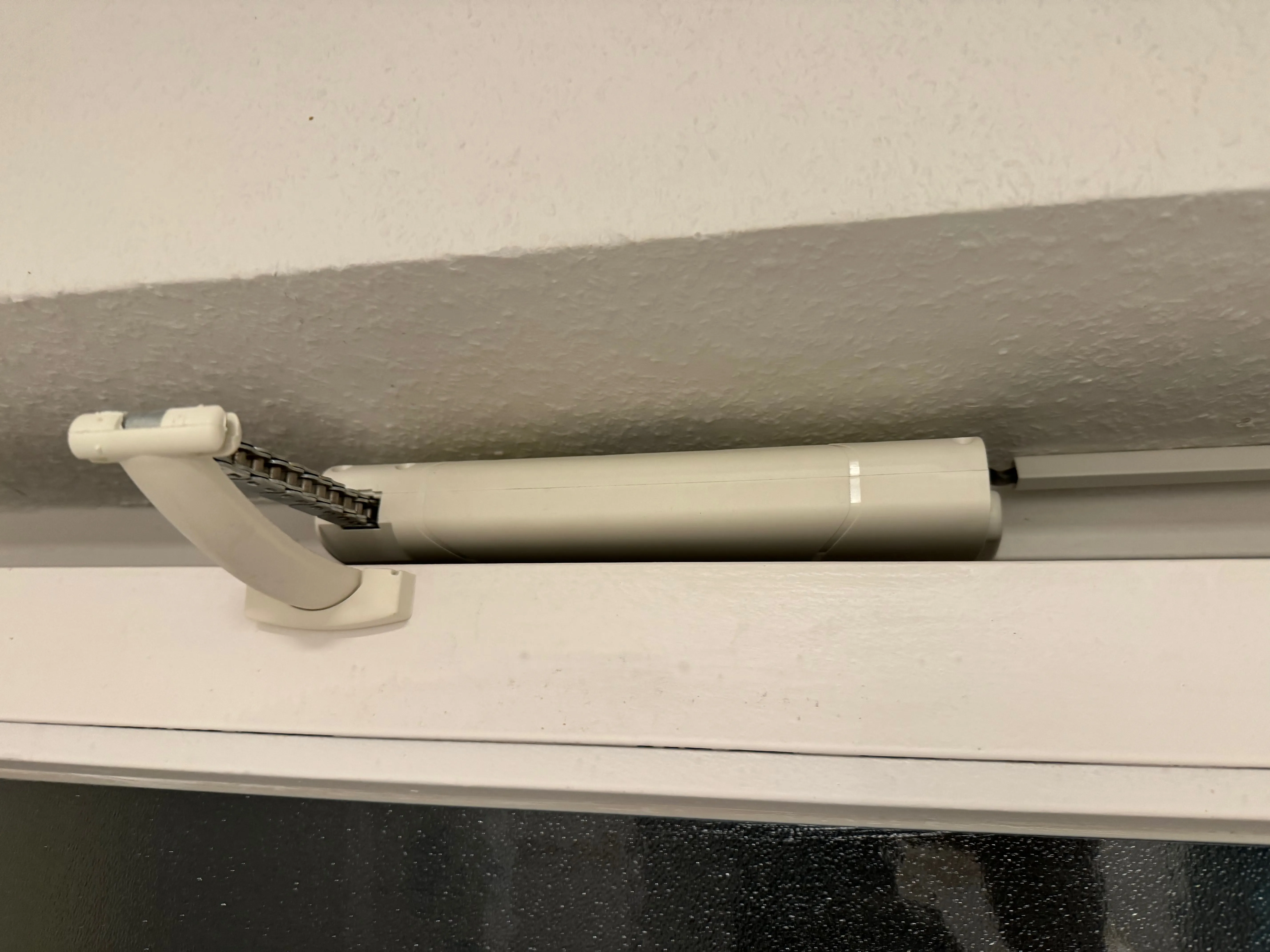 Close-up of a white cylindrical electric window opener motor mounted on a window frame, with a short section of black roller chain visible extending from the left end of the device into a pivoting bracket